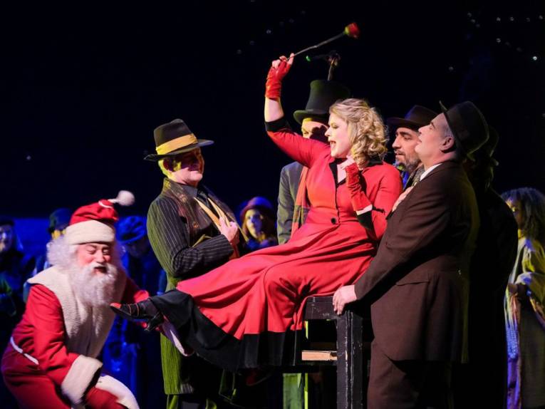 Vier Männer in Anzügen stehen um einen Stuhl, auf dem eine Frau in einem roten Mantel sitzt. Links im Bildhintergrund ist ein Mann in einem Weihnachtsmannkostüm zu sehen.